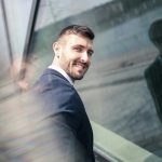 Businessman smiling confidently by a glass wall reflecting cityscape. Professional and optimistic vibe.